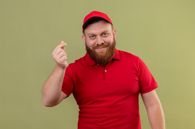 Giovane uomo di consegna barbuto in uniforme rossa e cappuccio che guarda l'obbiettivo sorridente strofinando le dita facendo gesto di denaro, in attesa di pagamento