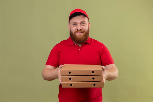 Giovane uomo di consegna barbuto in uniforme rossa e cappuccio che tiene pila di scatole per pizza sorridente fiducioso