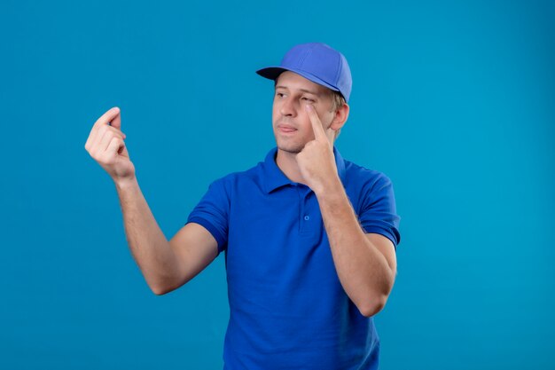 Giovane uomo di consegna bello in uniforme blu e berretto che osserva da parte indicando il suo occhio che fa gesto di soldi in attesa di un pagamento in piedi sopra la parete blu