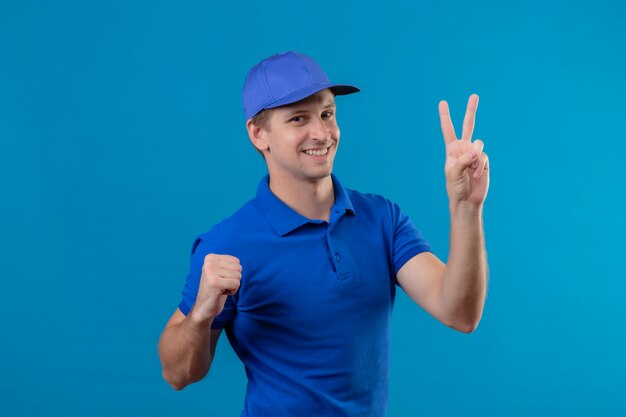 Giovane uomo di consegna bello in uniforme blu e cappuccio che mostra il numero due che sorride allegramente in piedi sopra la parete blu