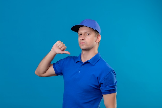 Giovane uomo di consegna bello in uniforme blu e cappuccio che punta a se stesso con il pollice che sembra fiducioso soddisfatto di sé e orgoglioso in piedi sopra la parete blu