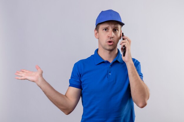 Giovane uomo di consegna bello in uniforme blu e cappuccio che sembra parlare confuso sul telefono cellulare