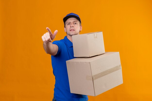 Giovane uomo di consegna bello in uniforme blu e cappuccio che tiene le scatole di cartone che indicano qualcosa con il dito