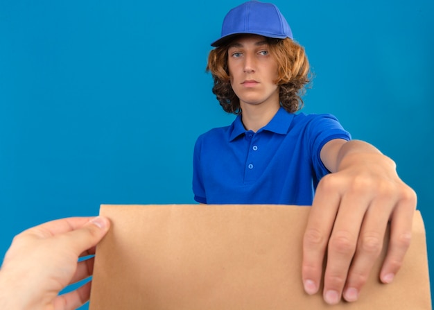 Giovane uomo di consegna che indossa la maglietta polo blu e cappuccio che dà un pacchetto di carta a un cliente con la faccia seria sopra fondo blu isolato