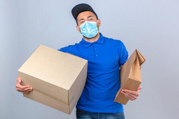 Giovane uomo di consegna che indossa la maglietta polo blu e il cappuccio nella mascherina medica protettiva in piedi con il pacchetto di carta e la scatola cercando fiduciosi su sfondo bianco isolato