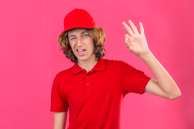 Giovane uomo di consegna che indossa la maglietta polo rossa e berretto che sbatte le palpebre guardando la fotocamera con un sorriso facendo segno ok in piedi su sfondo rosa isolato