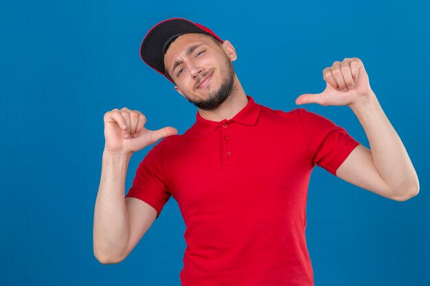 Giovane uomo di consegna che indossa la maglietta polo rossa e cappuccio guardando la telecamera sorridendo allegramente orgoglioso e soddisfatto di sé su sfondo blu isolato