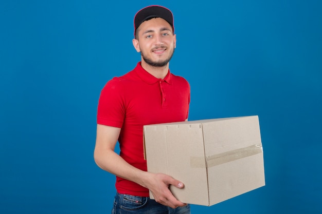 Giovane uomo di consegna che indossa la maglietta polo rossa e cappuccio in piedi con scatole di cartone guardando la fotocamera con un sorriso amichevole su sfondo blu isolato