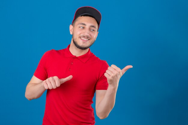 Giovane uomo di consegna che indossa maglietta polo rossa e cappello sorridente con la faccia felice guardando e indicando il lato con i pollici in su su sfondo blu isolato