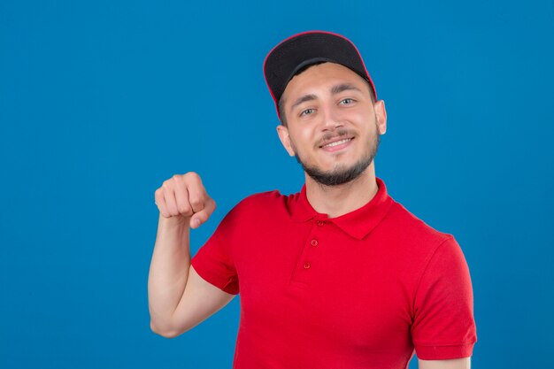 Giovane uomo di consegna che indossa maglietta polo rossa e cappuccio guardando la telecamera sorridendo amichevole gesticolando pugno bump come se il saluto di approvazione o come segno di rispetto isolato su sfondo blu