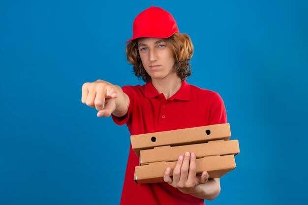 Giovane uomo di consegna che indossa uniforme rossa che tiene le scatole per pizza che punta dispiaciuto e frustrato per la fotocamera su sfondo blu isolato