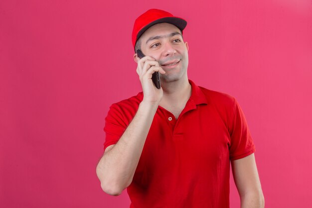 Giovane uomo di consegna in maglietta polo rossa e cappuccio parlando al telefono cellulare cercando soddisfatto su sfondo rosa isolato