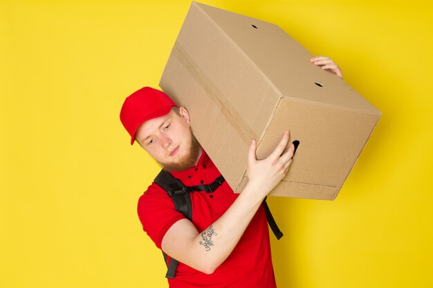 giovane uomo di consegna in polo bianco rosso cappello rosso jeans in possesso di una scatola su giallo