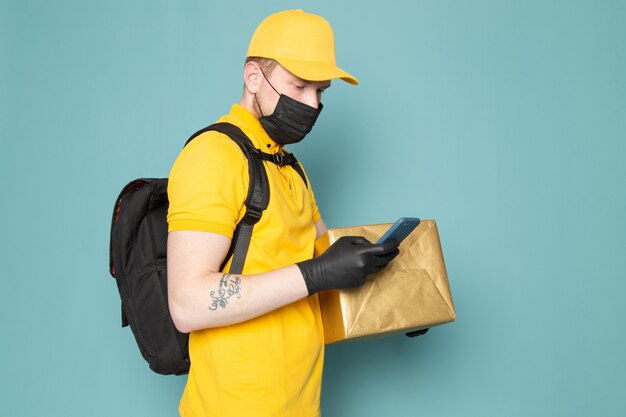 giovane uomo di consegna in polo giallo berretto giallo jeans bianchi zaino e maschera sterile nera in possesso di una scatola su blu