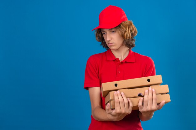 Giovane uomo di consegna in rosso uniforme tenendo le scatole per pizza guardando da parte con espressione triste su sfondo blu isolato