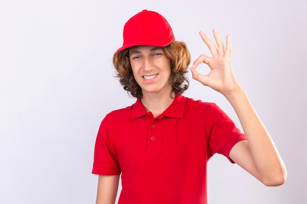 Giovane uomo di consegna in uniforme rossa che guarda l'obbiettivo sorridente amichevole facendo segno ok su sfondo bianco isolato