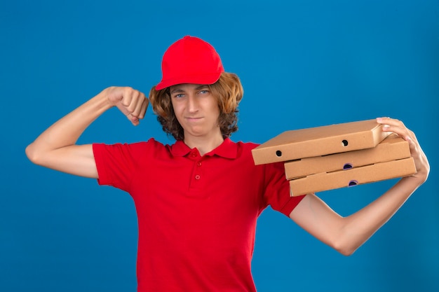 Giovane uomo di consegna in uniforme rossa in piedi con scatole per pizza sulla spalla alzando il pugno cercando fiducioso concetto vincitore su sfondo blu isolato