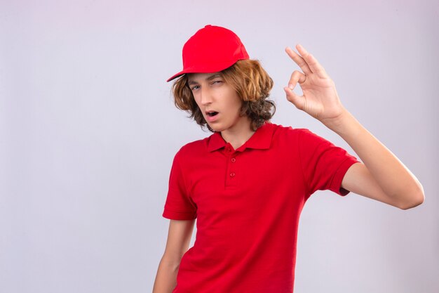 Giovane uomo di consegna in uniforme rossa sorridente allegramente facendo segno ok su sfondo bianco isolato su sfondo isolato