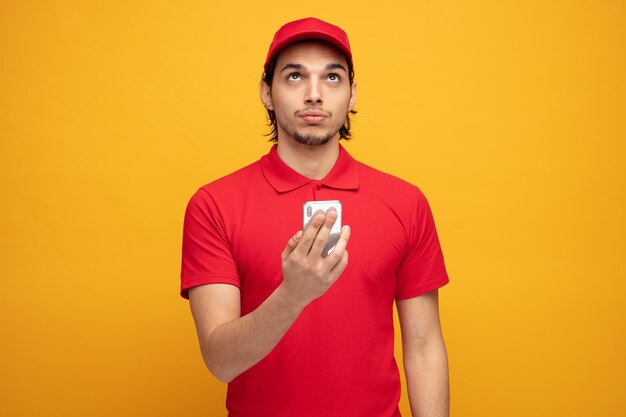 giovane uomo di consegna serio che indossa l'uniforme e il cappuccio che tiene il telefono cellulare guardando in alto isolato su sfondo giallo