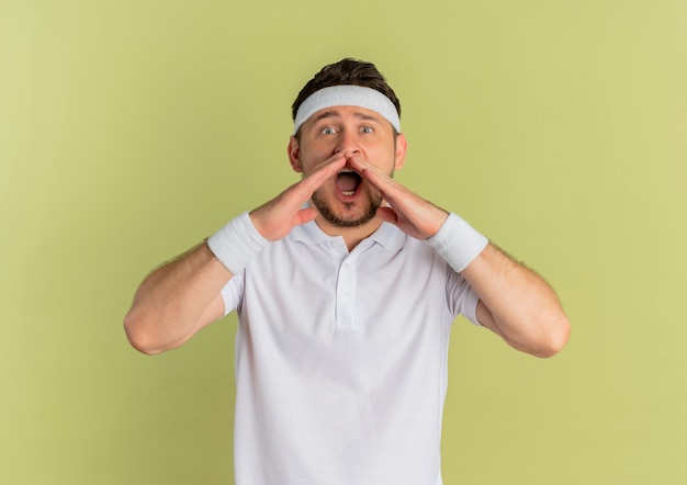 Giovane uomo di forma fisica in camicia bianca con fascia che grida in preda al panico con le mani vicino alla bocca in piedi sopra il muro di oliva