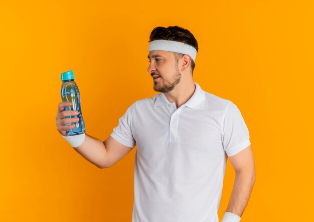 Giovane uomo di forma fisica in camicia bianca con la fascia tenendo la bottiglia di acqua guardandolo in piedi su sfondo arancione