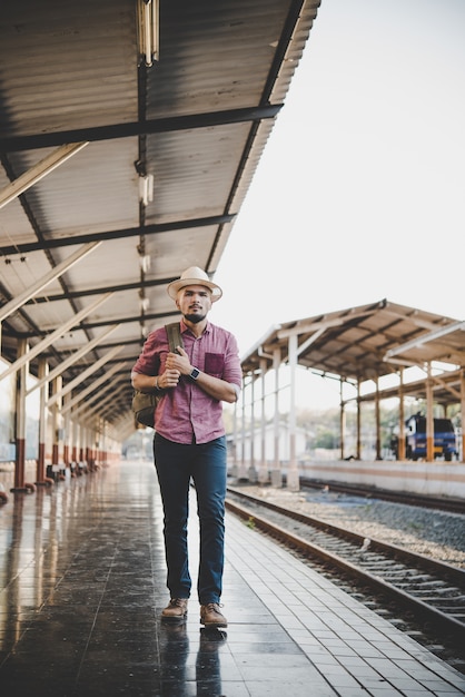 Giovane uomo di hipster che cammina attraverso la stazione ferroviaria. L&#39;uomo in attesa del treno sulla piattaforma. Concetto di viaggio.