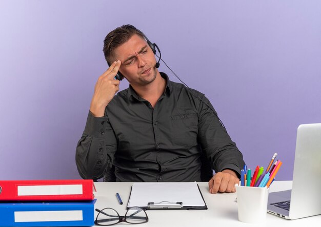 Giovane uomo di lavoratore di ufficio bionda infastidito sulle cuffie si siede alla scrivania con strumenti di office guardando il laptop mette la mano sulla testa isolata su sfondo viola con spazio di copia