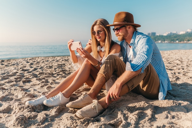 Giovane uomo e donna felici sorridenti attraenti in occhiali da sole che si siedono sulla spiaggia della sabbia che prende la foto del selfie