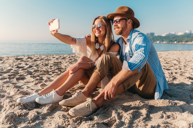 Giovane uomo e donna felici sorridenti attraenti in occhiali da sole che si siedono sulla spiaggia della sabbia che prende la foto del selfie