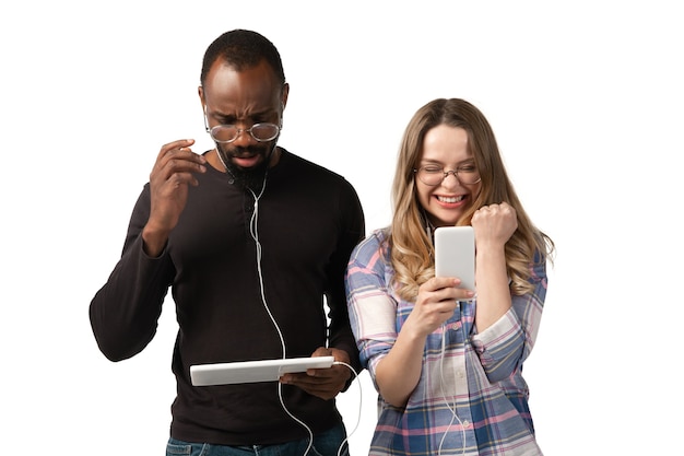 Giovane uomo e donna utilizzando laptop, dispositivi, gadget isolati sulla parete bianca dello studio