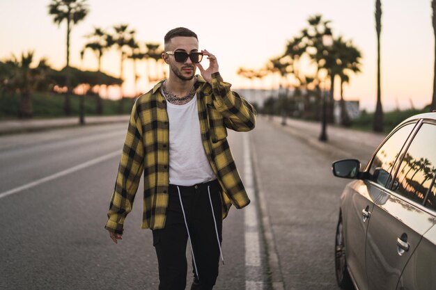 Giovane uomo europeo alla moda in una camicia a quadri e occhiali da sole che cammina per strada