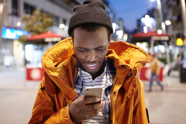 Giovane uomo europeo nero attraente in messaggio di testo di battitura a macchina dell'abbigliamento di inverno sul suo cellulare, stante nella regolazione della città di notte. SMS di lettura maschio dalla pelle scura gioiosa