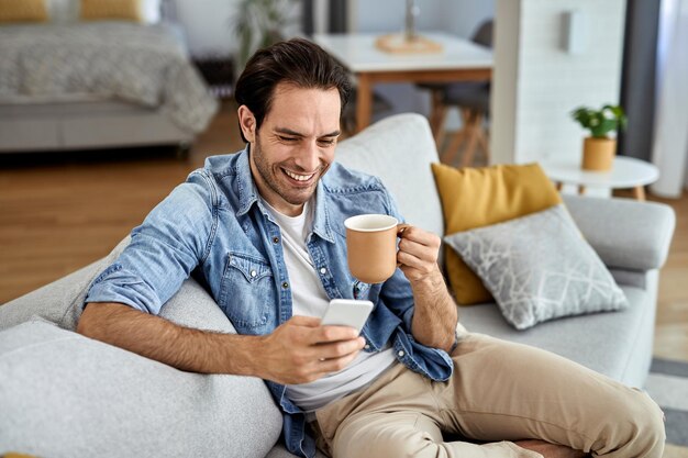 Giovane uomo felice che manda un sms sullo smartphone mentre ci si rilassa sul divano e bevendo un caffè