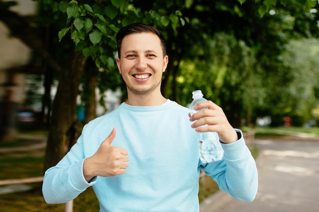 Giovane uomo felice con la bottiglia di acqua nella sua mano che sorride e che mostra i pollici su