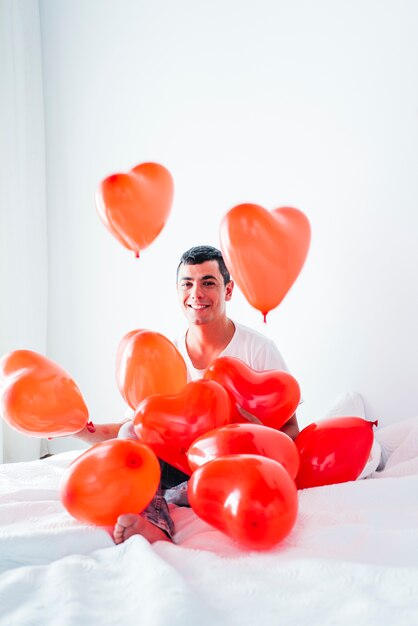 Giovane uomo felice sul letto tra palloncini a forma di cuori