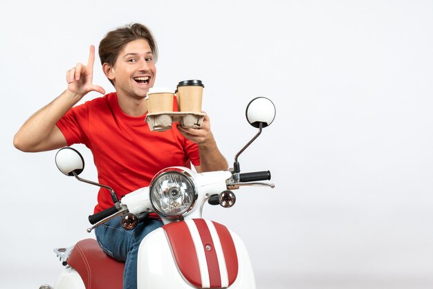 Giovane uomo fiducioso del corriere in uniforme rossa che si siede sullo scooter e che tiene gli ordini sulla parete gialla