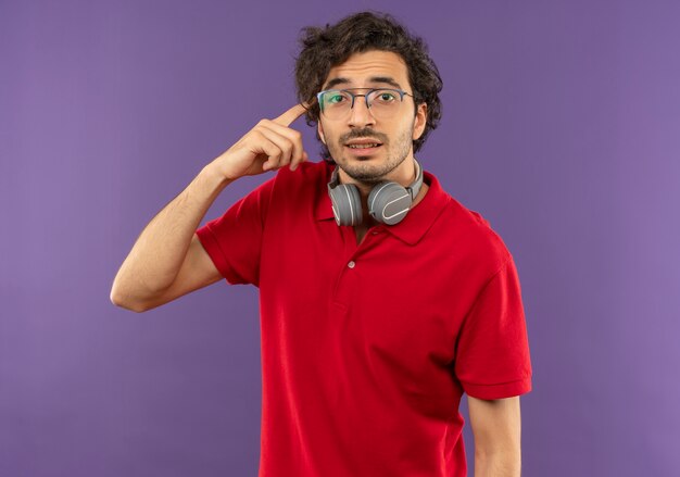 Giovane uomo fiducioso in camicia rossa con occhiali ottici e con le cuffie mette il dito sulla testa isolata sulla parete viola