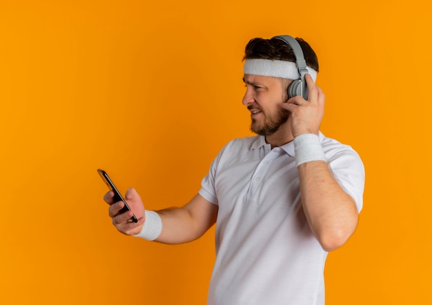 Giovane uomo fitness in camicia bianca con archetto e cuffie guardando lo schermo del suo cellulare alla ricerca di musica in piedi su sfondo arancione