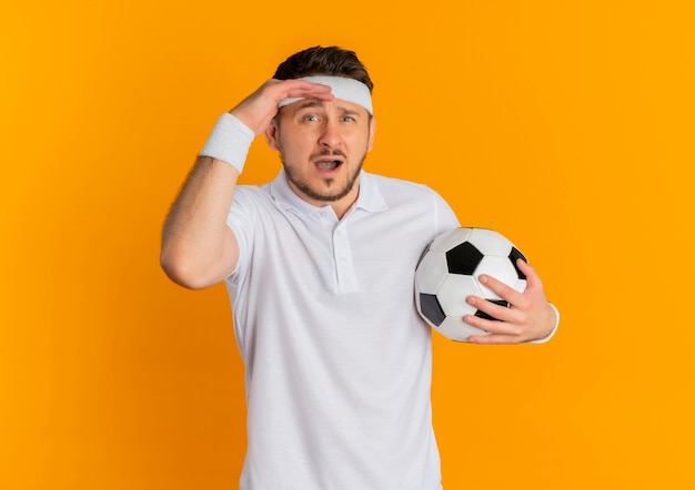 Giovane uomo fitness in camicia bianca con fascia tenendo il pallone da calcio guardando la telecamera confusa in piedi su sfondo arancione