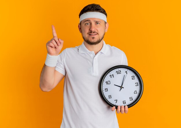 Giovane uomo fitness in camicia bianca con fascia tenendo l'orologio da parete rivolto verso l'alto con il dito guardando la fotocamera con il sorriso sul viso in piedi su sfondo arancione