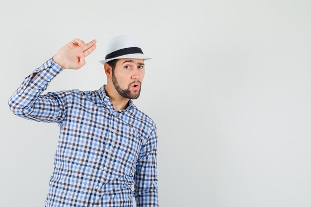 Giovane uomo gesticolando con la mano e le dita in camicia a quadri, cappello e guardando fiducioso, vista frontale.