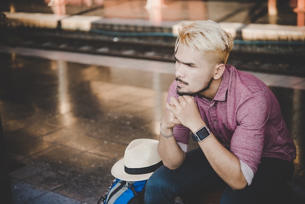 Giovane uomo hipster seduto sulla panca di legno con zaino alla stazione ferroviaria. Uomo seduto in attesa del treno alla piattaforma.