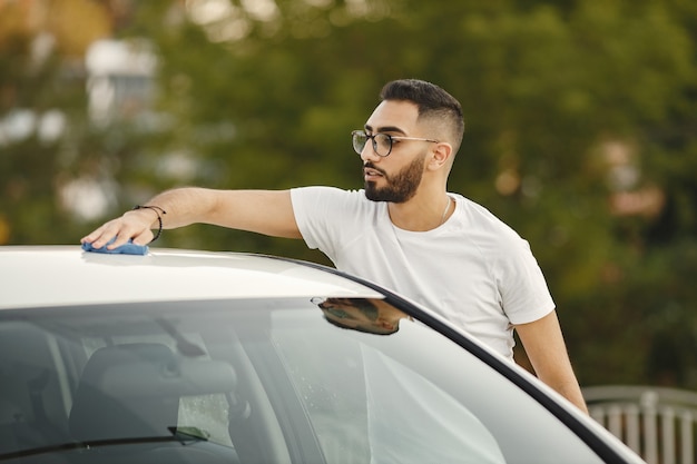 Giovane uomo in abiti alla moda che pulisce la sua auto con un tappeto dopo il lavaggio alla stazione di autolavaggio. Uomo che indossa una maglietta bianca e jeans