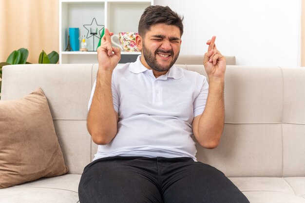 Giovane uomo in abiti casual che esprime il desiderio desiderabile incrociando le dita con l'espressione di speranza con gli occhi chiusi seduto su un divano in un soggiorno luminoso