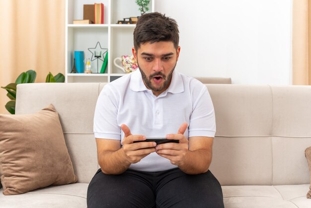 Giovane uomo in abiti casual che gioca usando lo smartphone emotivo e sorpreso seduto su un divano in un soggiorno luminoso