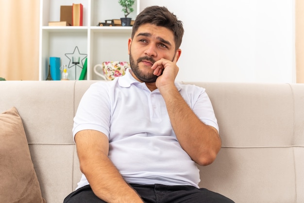 Giovane uomo in abiti casual che guarda da parte perplesso seduto su un divano in un soggiorno luminoso