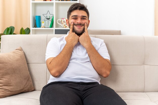 Giovane uomo in abiti casual che guarda indicando con l'indice il suo sorriso falso seduto su un divano in un soggiorno luminoso
