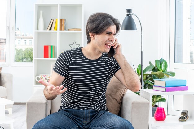 Giovane uomo in abiti casual che sembra confuso e dispiaciuto mentre parla al telefono cellulare seduto sulla sedia in un soggiorno luminoso