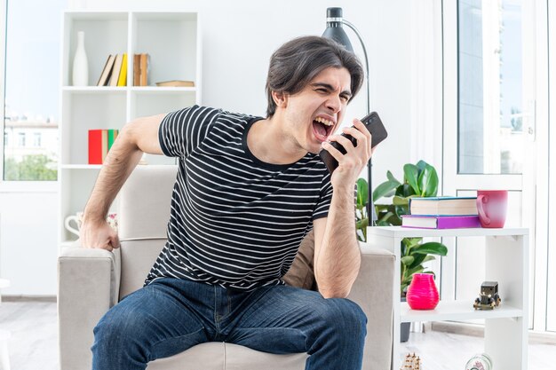 Giovane uomo in abiti casual che sembra eccitato e frustrato che grida mentre parla al telefono cellulare seduto sulla sedia in un soggiorno luminoso