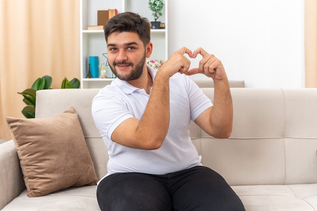 Giovane uomo in abiti casual che sembra felice e positivo che fa un gesto di cuore con le dita seduto su un divano in un soggiorno luminoso